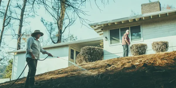 Home ignition zone assessment: Watering dry brush near house for wildfire safety.